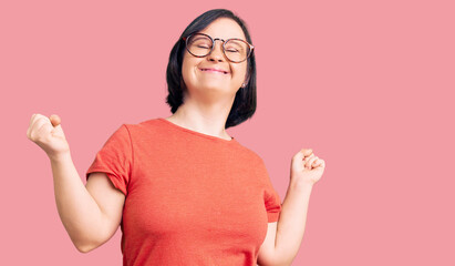 Brunette woman with down syndrome wearing casual clothes and glasses very happy and excited doing winner gesture with arms raised, smiling and screaming for success. celebration concept.