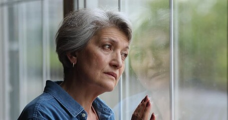 Close up view sad older 60s woman standing indoor near window look outside feels depressed, quarantine self-isolation time due covid pandemic, goes through divorce, loss of loved one, nostalgy concept
