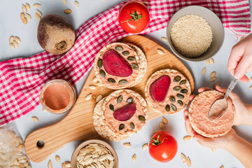 Crispy puffed rice cakes on table hummus spread and tomato and beet vegetables and sesame pumpkin seeds on the table - top view on hands preparing healthy vegetarian or vegan breakfast gluten free