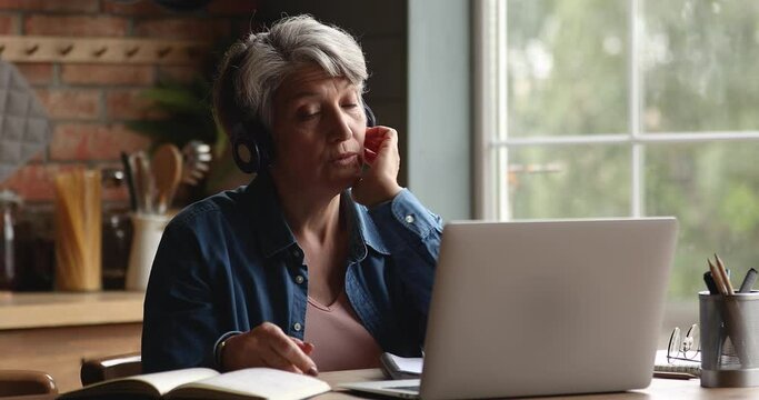 Aged woman wear headphones talk to family by videocall app on laptop. E-learn with online tutor, videoconference event, gain new knowledge distantly for new opportunities earning on retirement concept