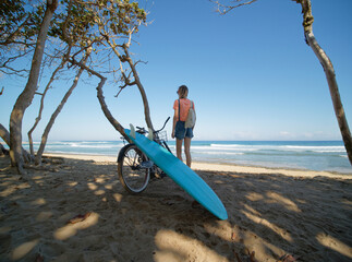 Surfer Girl Stands With Bicycle On The Beach With Surfboard Checks Waves in Ocean. Rear View....
