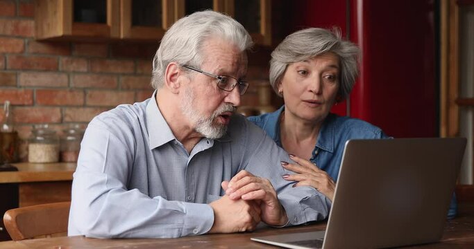 Grey haired elderly couple sit in kitchen at home using laptop app talk by videocall. Communication distantly due covid-19 quarantine, on-line counselling, Video call event, modern technology concept