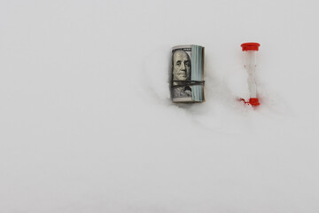 A dollar in winter, a roll of rolled dollar bills lying on the snow with a red hourglass, close-up, the concept of time making money.