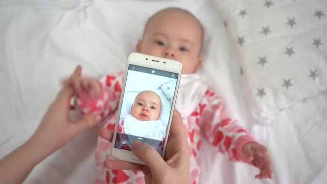 Mother standing near the cradle makes photos with the baby on the phone.