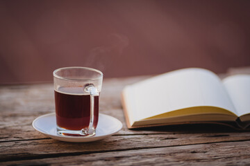 A cup of hot morning coffee on wooden table with a diary notebook at the balcony of the sleeping room to welcome sunshine in the morning, a glass of black coffee with a notebook on the wooden table