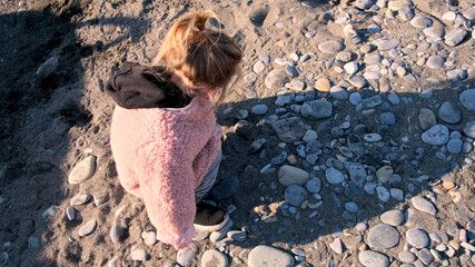 Blonde little girl with pigtail in fluffy pink jacket plays with sand and stones on sea beach at autumn sunset close upper view