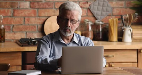 Grey-haired single male sit in kitchen wear glasses using laptop device feels puzzled over internet application or new software, older man experiences difficulties with modern technology usage concept - Powered by Adobe
