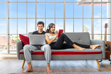 Young Asian couple relaxing sitting on sofa using laptop computer and digital tablet working from home
