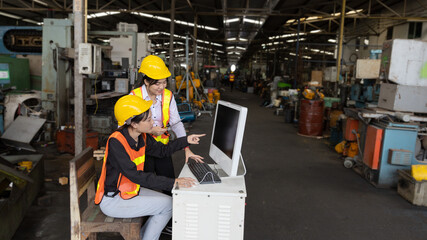 Two factory workers or technicians are using computer and technology to check stock in a heavy machinery warehouse