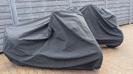 two motorcycles protected by black grey protective cover in city street motorbike with tarpaulin...