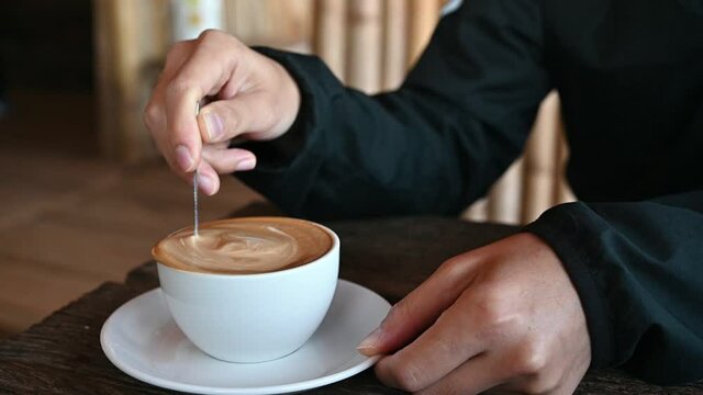 Someone hand stirring hot latte coffee with spoon before drinking. A latte is a coffee drink made with espresso and steamed milk.