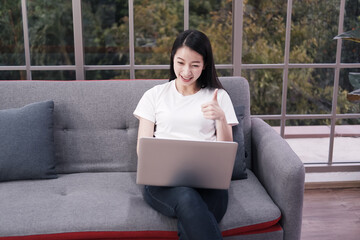 Smiling Asian businesswoman in white shirt is sitting on sofa and online working with laptop at home. She is blogger freelance in social media website. Communication and technology concept.