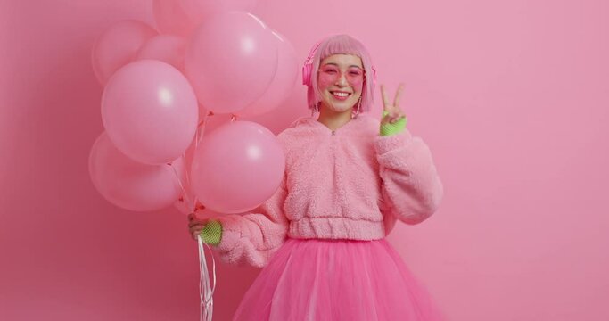 Happy delighted young Asian woman with pink hair makes victory gesture smiles pleasantly dressed in fashionable festive clothes for party holds inflated balloons enjoys anniversary celebration
