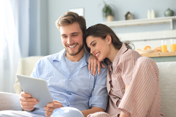 Young couple in pajamas watching media content online in a tablet sitting on a sofa in the living room.