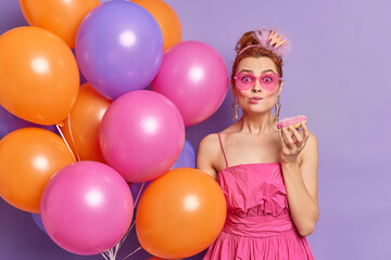 Fototapeta na wymiar Pretty redhead woman holds delicious sweet doughnut eats tasty dessert on party poses with inflated balloons in fashionable clothes isolated over purple background. Birthday celebration concept