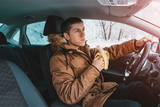 Male Driver Has A Sharp Drop In Blood Sugar And He Urgently Eats A Banana While Driving Through The Streets In Winter. The Concept Of Fast Carbs