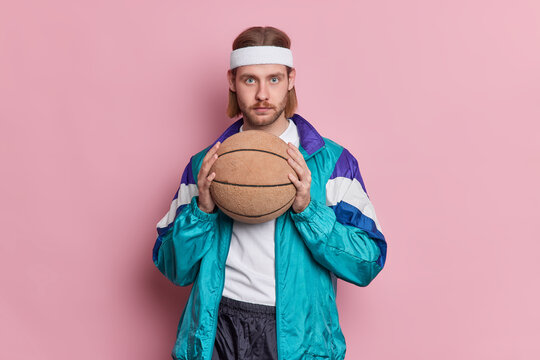 Serious Blue Eyed Male Basketball Player With Stubble Long Hair Holds Ball Ready For Playing Game Wears White Headband And Sportsclothes Poses Against Pink Background. Active Lifestyle Concept