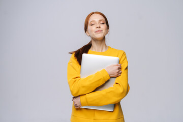 Obraz premium Happy charming young woman student holding laptop computer with closed eyes on isolated gray background. Pretty lady model with red hair emotionally showing facial expressions in studio, copy space.