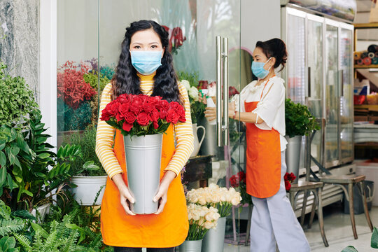 Flower shop owner with bucket of roses and her colleague in medical masks getting ready to open the shop in the morning