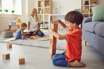 Cute little boy building wood block tower with small brother and mommy in background