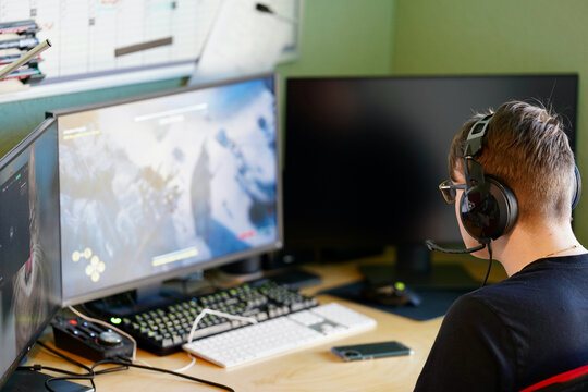Young man playing computer games with joystick, headset on and three monitors.