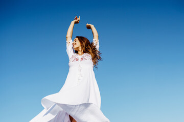 Young woman raising her arms in a beautiful white dress against a blue sky