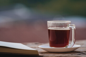 A cup of hot morning coffee on wooden table with a diary notebook at the balcony of the sleeping room to welcome sunshine in the morning, a glass of black coffee with a notebook on the wooden table