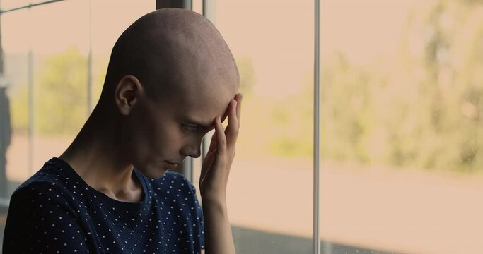 Side close up view portrait of bald young female cancer patient standing indoor bowing her shaved head looking out the window thinking about illness feels hopeless. Oncology disease relapse concept