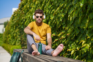Male sitting outside using an aluminum water bottle, headphones and backpack.