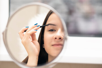 A young woman combs her eyebrows with a brush while looking in the mirror