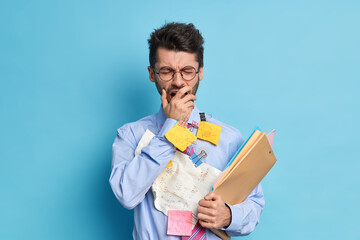 Stressed student has much work to do yawns after sleepless night has tired expression dressed in elegant shirt carries papers makes memo stickers to remember mathematical formulas. Studying concept