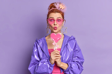 Beautiful surprised woman with red hair dressed in retro style wears sunglasses jacket and big earrings holds heart shaped lollipop keeps lips rounded poses against purple background. Nineties style.