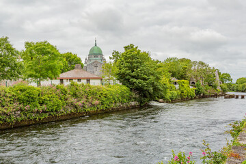 Fototapeta na wymiar River Corrib with its calm waters surrounded by green vegetation and the cathedral with its green dome in the background, Waterways of Galway, cloudy day in Galway city, Connacht province, Ireland