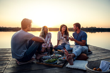 Group of friends having fun on picnic near a lake, sitting on pier eating and drinking wine.