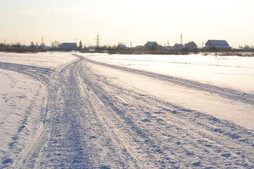Fototapeta premium Winter bright landscape. Snowmobile tracks in the snow in the middle of snowdrifts in a snow-covered field against the backdrop of the bright winter sun and silhouettes of village houses.