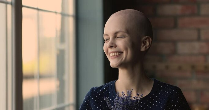 Head shot happy bald female cancer patient with lavender flowers bunch pose indoor smiles looks at camera feel alive, victory over oncology disease, remission, full recovery. World Cancer Day concept