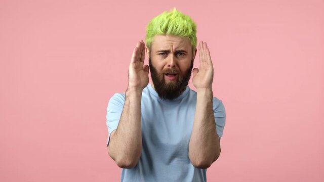 Overemotional bearded man freak with dyed bright green hair closing face with palms of hands demonstrating different emotions, hiding his personality. Indoor studio shot isolated on pink background