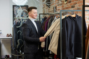 A young stylish man in a suit is choosing a shirt or hoodie in a clothing store. A male customer in a boutique.