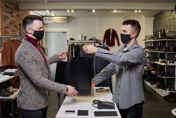 A man in a face mask to avoid the spread of coronavirus is taking his purchase from a seller in a clothing store. A male shop assistant is giving a second bag with clothes to a customer in a boutique