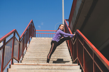 young athletic girl stands with her foot on the railing of the stairs, workout urban sport concept