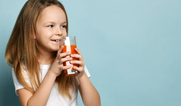 Cheerful happy small cute girl in white t-shirt standing with glass of fresh fruit juice and looking aside over blue background, copy space. Happy childhood, healthy lifestyle concept