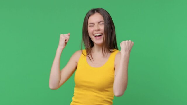 Excited overjoyed brunette young woman 20s in yellow basic tank top posing isolated on green background in studio. People lifestyle concept. Doing winner gesture celebrating clenching fists say yes