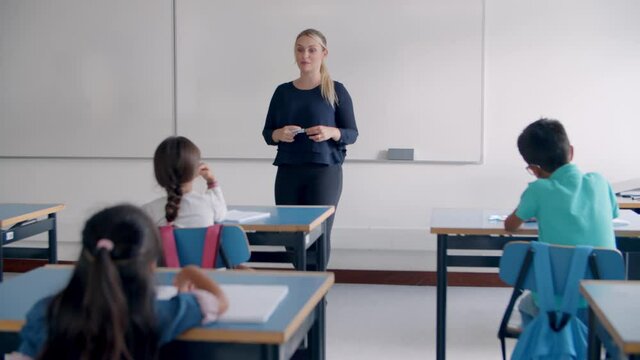 Female teacher giving and explaining task to pupils, asking questions in class. Students raising hands at desks. Teaching or primary education concept