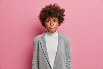 Photo of nervous puzzled dark skinned young businesswoman presses lips and looks embarrassed at camera wears round transparent spectacles dressed in elegant clothes isolated over pink background