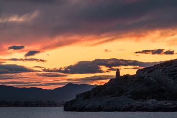 Atardecer con cielo naranja con nubes, montañas de fondo y el relieve de un faro el la costa...
