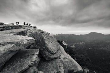 Fotografia en blanco y negro de una montaña rocosa con gente en la cumbre en un dia de nubes