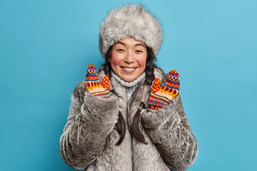 Pretty Eskimo woman dresses for cold weather smiles happily wears fur coat and hat raises hands in mittens has good mood isolated over blue background. Asian female from tundra in winter clothes. - obrazy, fototapety, plakaty