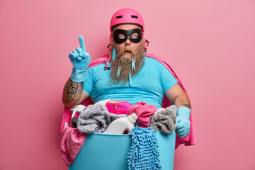 Suprised overweight superhero in helmet tsirt and rubber gloves stands with basin of dirty linen...