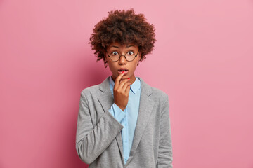 Fototapeta na wymiar Stupefied African American female manager in formal outfit keeps mouth opened stares surprised at camera wears round spectacles isolated over pink background. People bussiness face expressions