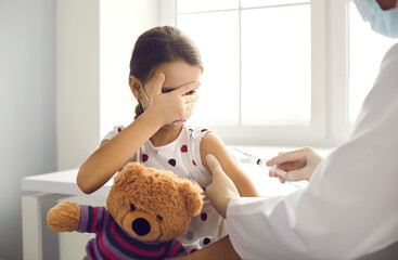 Little kid terrified by injection at the hospital. Girl afraid of syringe needle covers face while getting flu vaccine at pediatric clinic. Doctor giving a shot to scared child holding teddy bear toy - obrazy, fototapety, plakaty